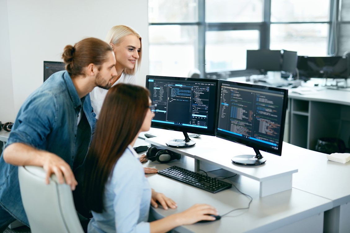 Group of three IT employees working on a program at the office using two monitors